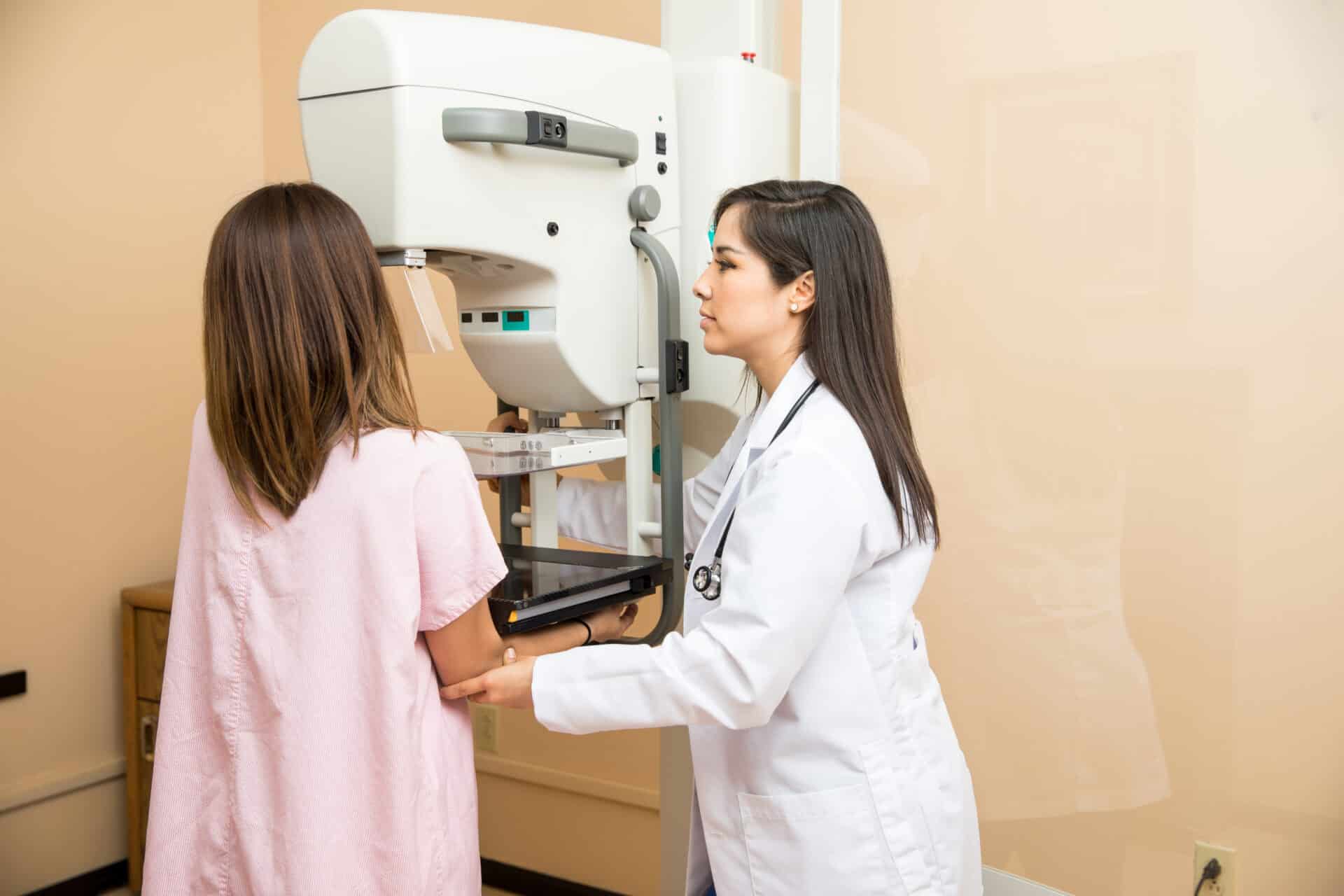 A healthcare professional assists a woman undergoing a mammogram. The patient stands in front of a mammography machine, wearing a pink gown, while the medical staff, in a white coat, positions her for the procedure.