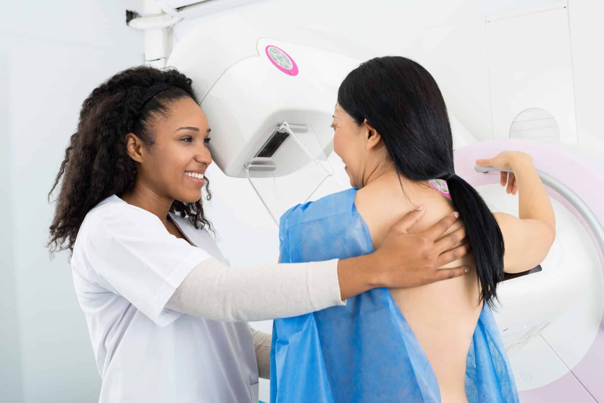 A healthcare professional assists a patient with dark hair during a mammogram. The patient is partially covered with a blue gown and standing in front of the mammography machine. Both are smiling, creating a supportive atmosphere.