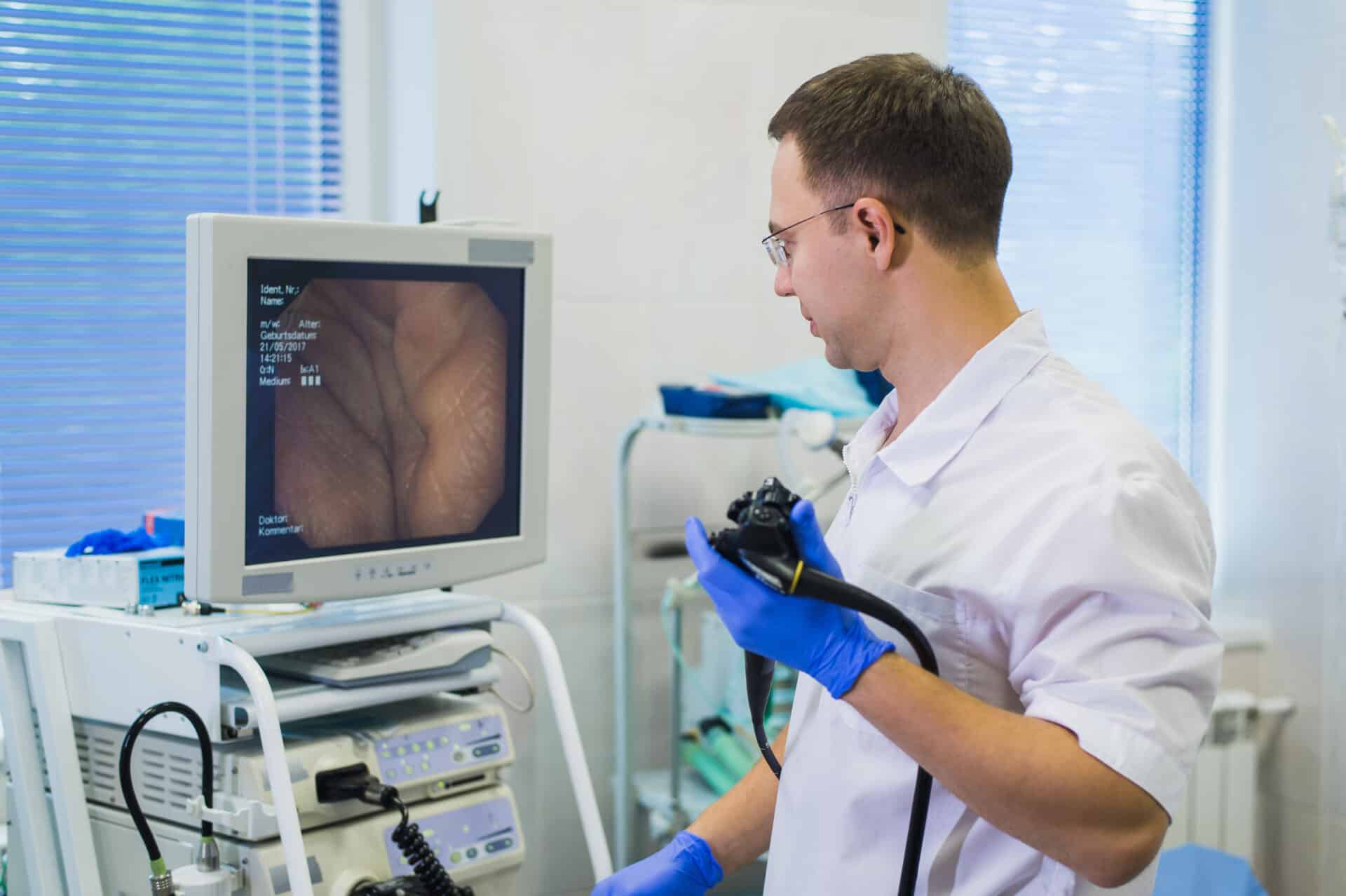 A medical professional wearing gloves holds an endoscope and looks at a monitor displaying an endoscopic image. The room contains medical equipment, creating a professional healthcare setting.