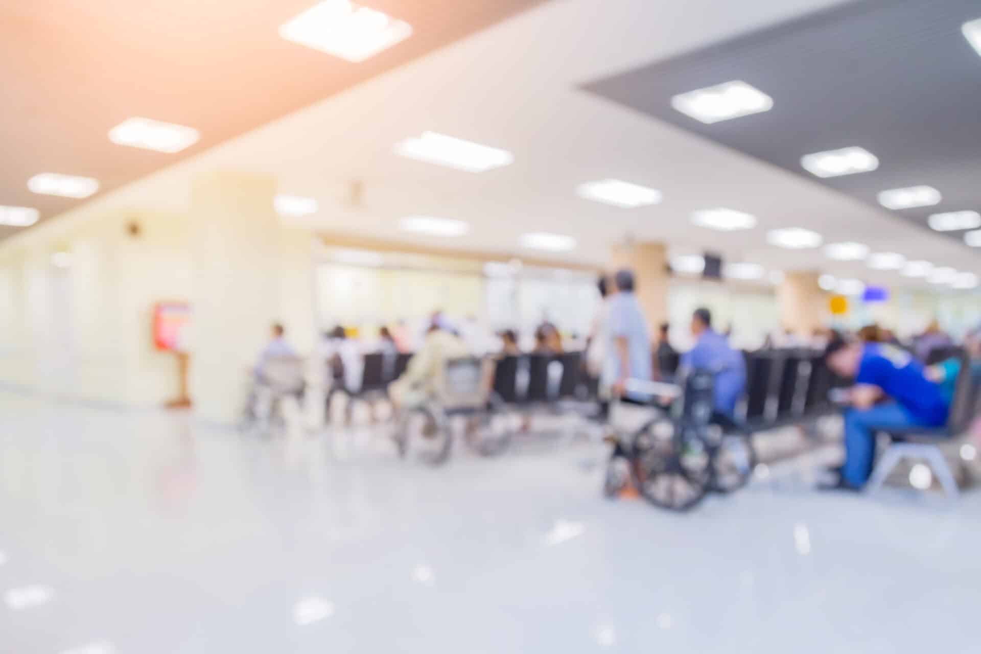 A blurred image of a hospital waiting area with people sitting on chairs, some in wheelchairs. The lighting is bright and the floor appears shiny, reflecting the overhead lights.