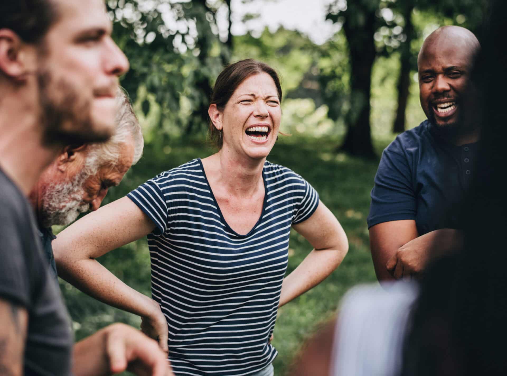 A group of people outdoors are laughing and enjoying each other's company. A woman in a striped shirt stands at the center, smiling broadly. Surrounding trees and greenery enhance the relaxed, joyful atmosphere.