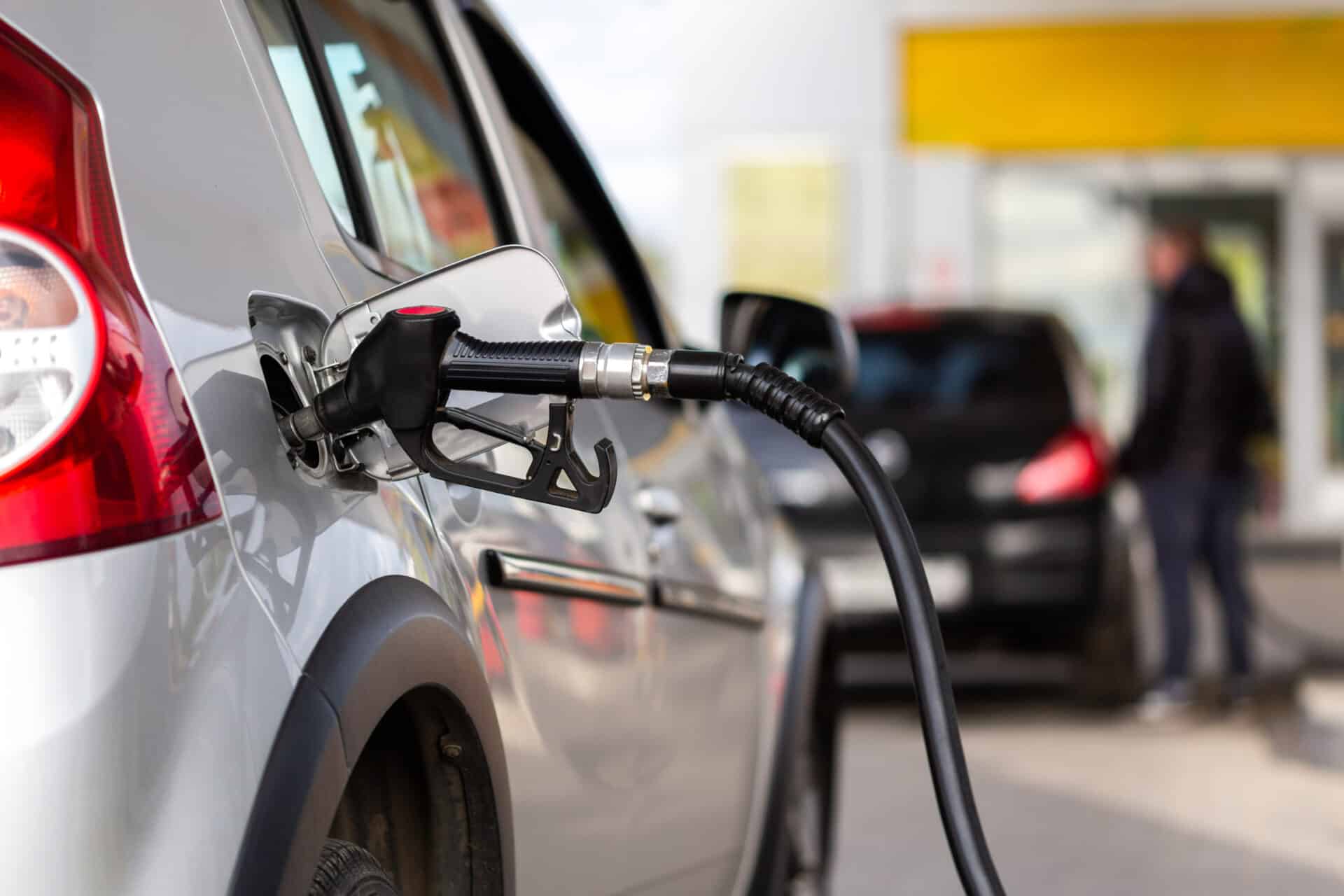 A silver car is being refueled at a gas station. The fuel nozzle is inserted into the car's gas tank. In the background, a person stands near another car. The station has a yellow and white canopy.