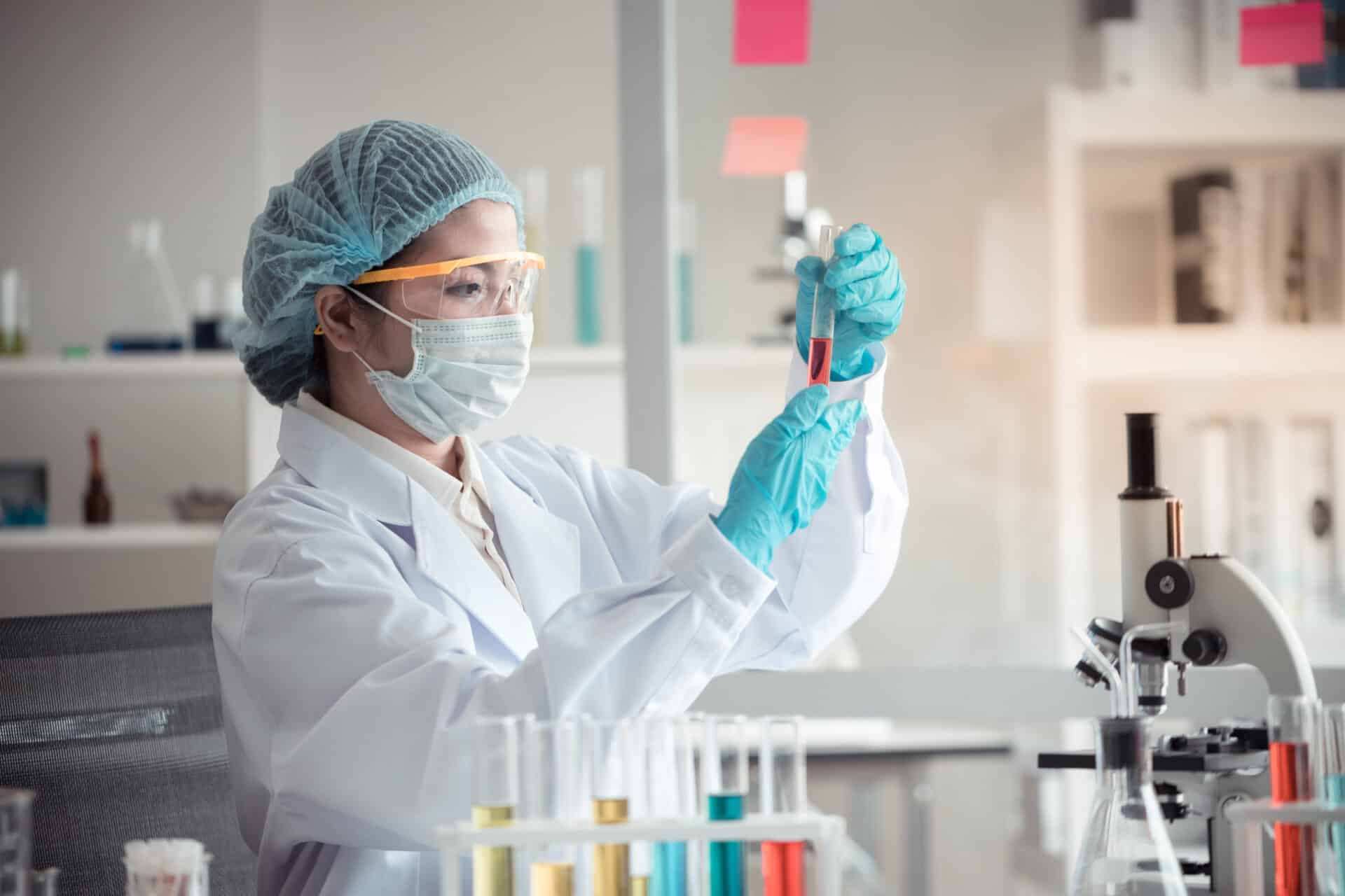 A scientist in protective gear, including a lab coat, mask, goggles, and gloves, examines a test tube containing a red liquid. They are in a laboratory setting with equipment and test tubes visible in the background.