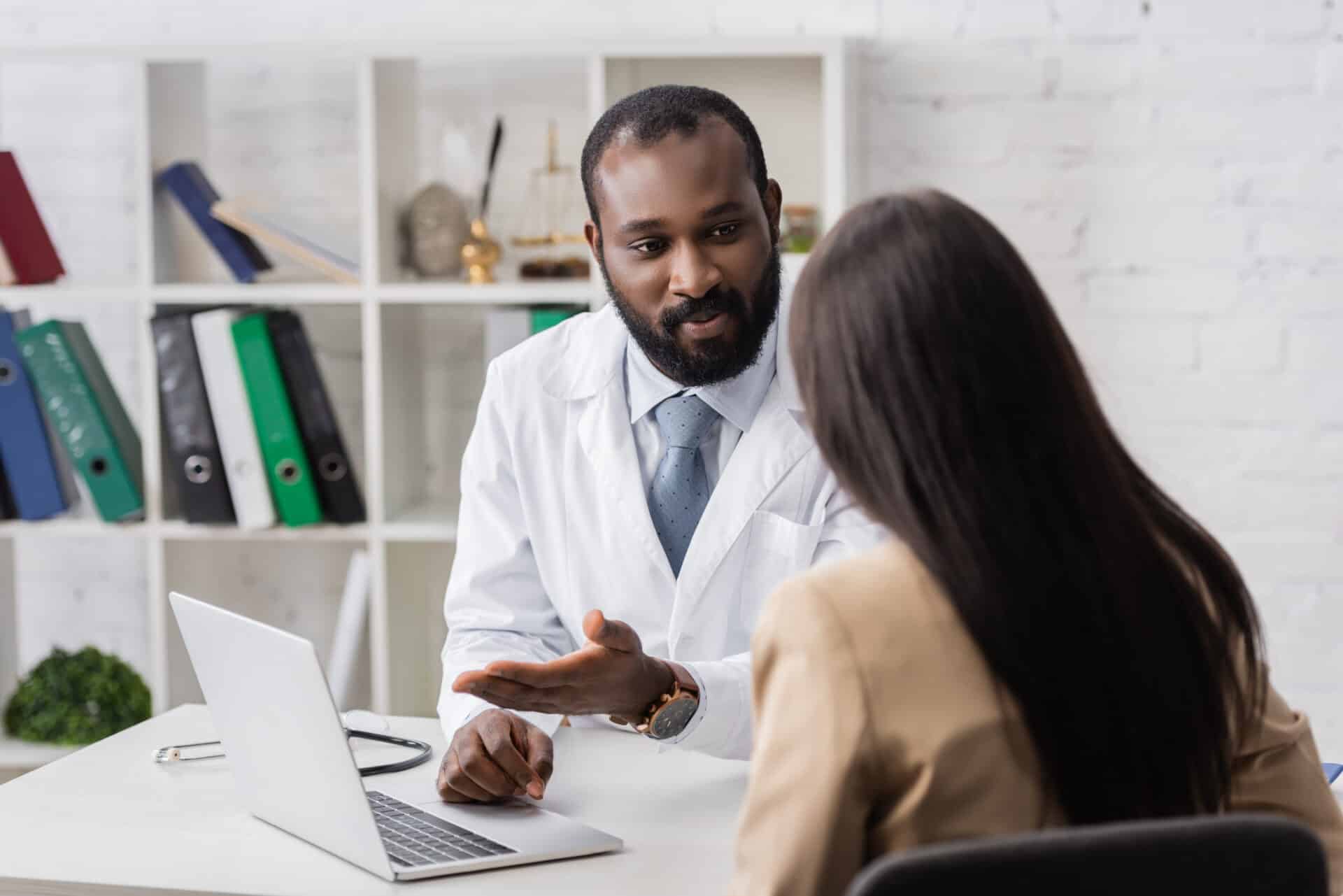 A doctor in a white coat sits at a desk, engaging in conversation with a patient. The doctor gestures with one hand. A stethoscope and a laptop are on the desk. Shelves with books and binders are in the background.