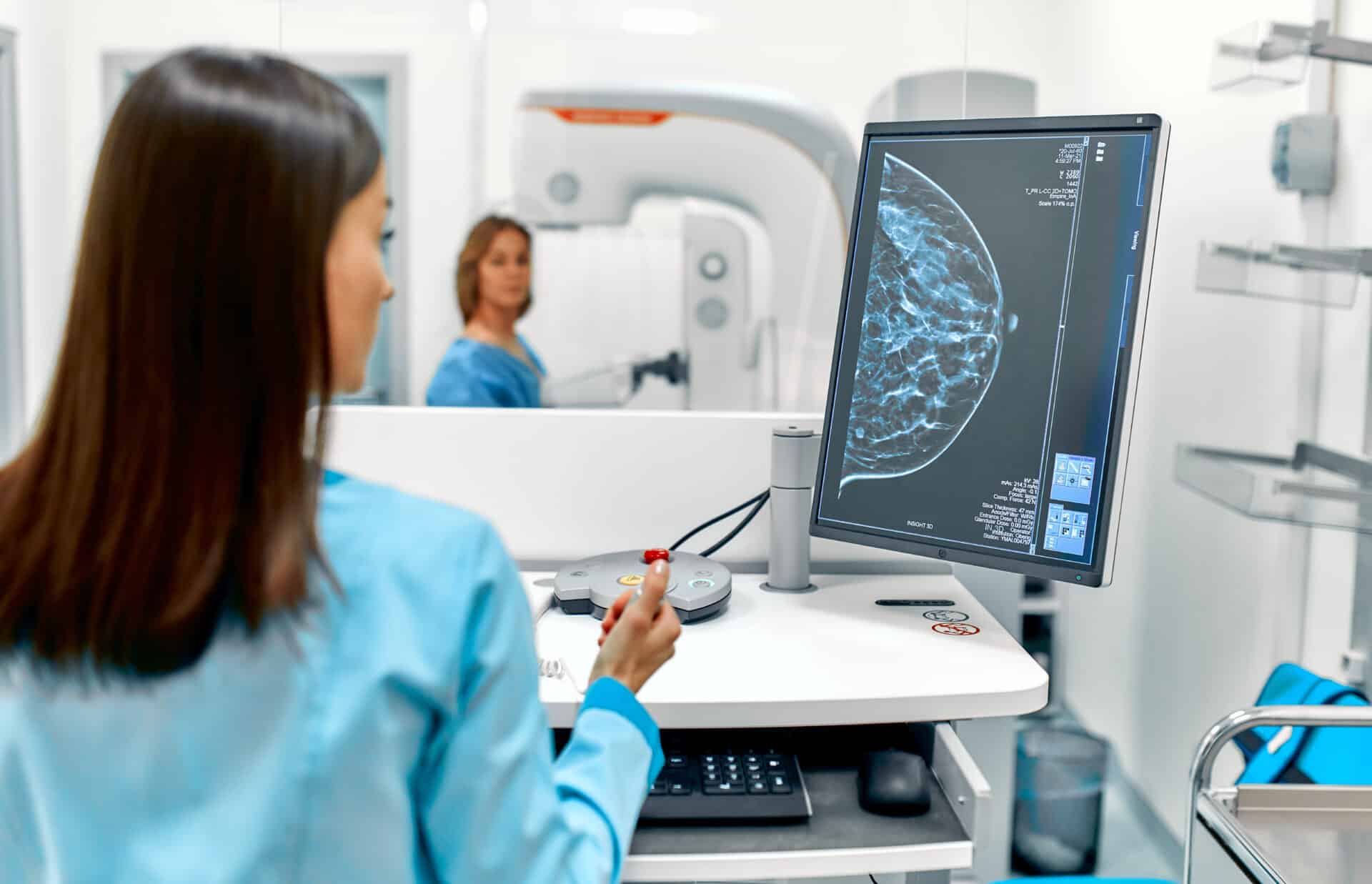 A healthcare professional examines a mammogram image on a computer screen in a medical office. A patient stands near a mammography machine in the background, with medical equipment visible around the room.