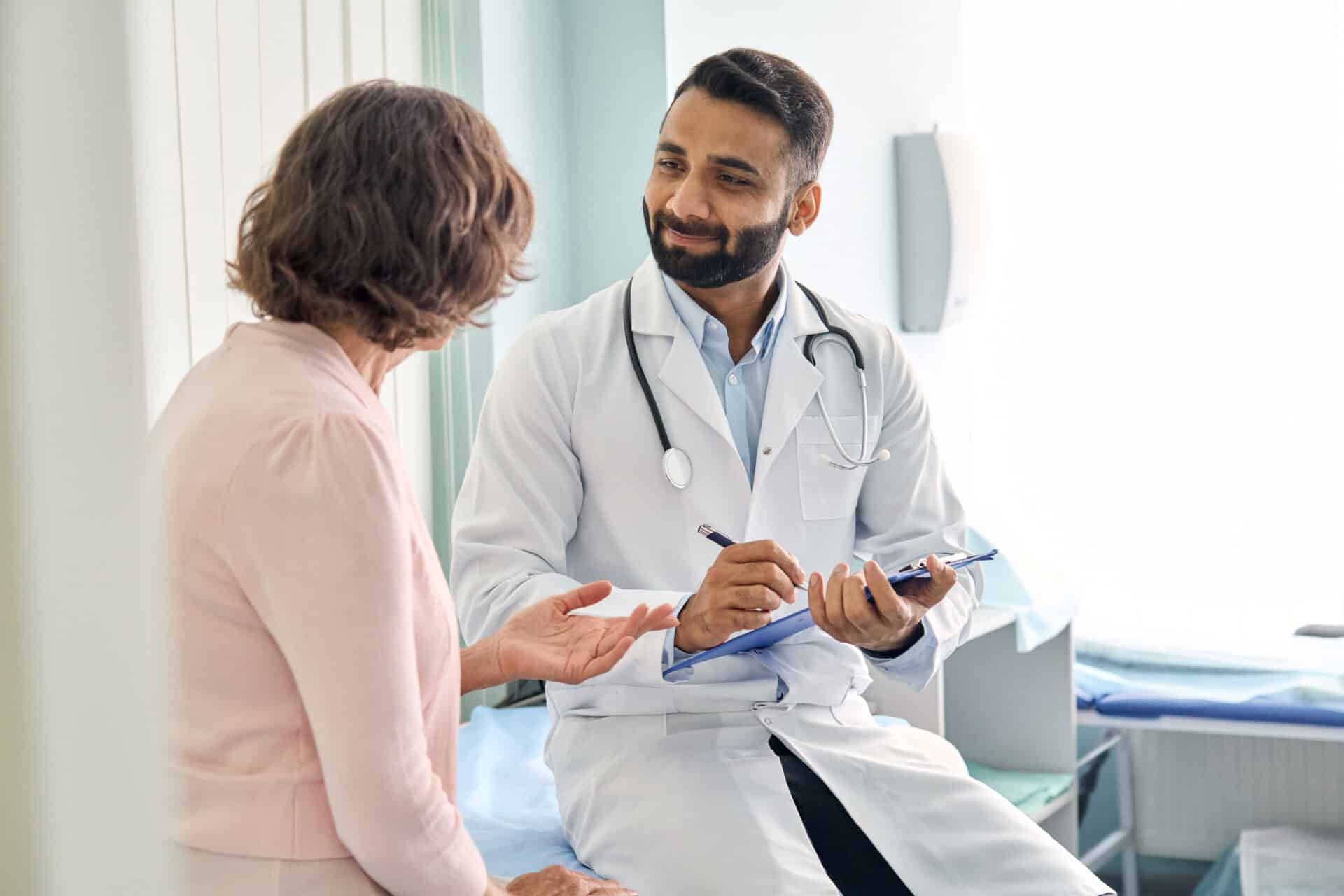 A doctor in a white coat with a stethoscope sits and listens attentively to a patient, holding a clipboard. The patient gestures with her hands while speaking in a bright medical office.