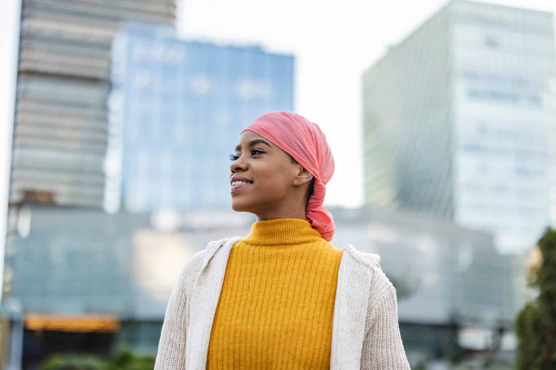 A person wearing a yellow sweater and a pink headscarf smiles while standing outdoors. They are in an urban area with modern glass buildings in the background on a clear day.