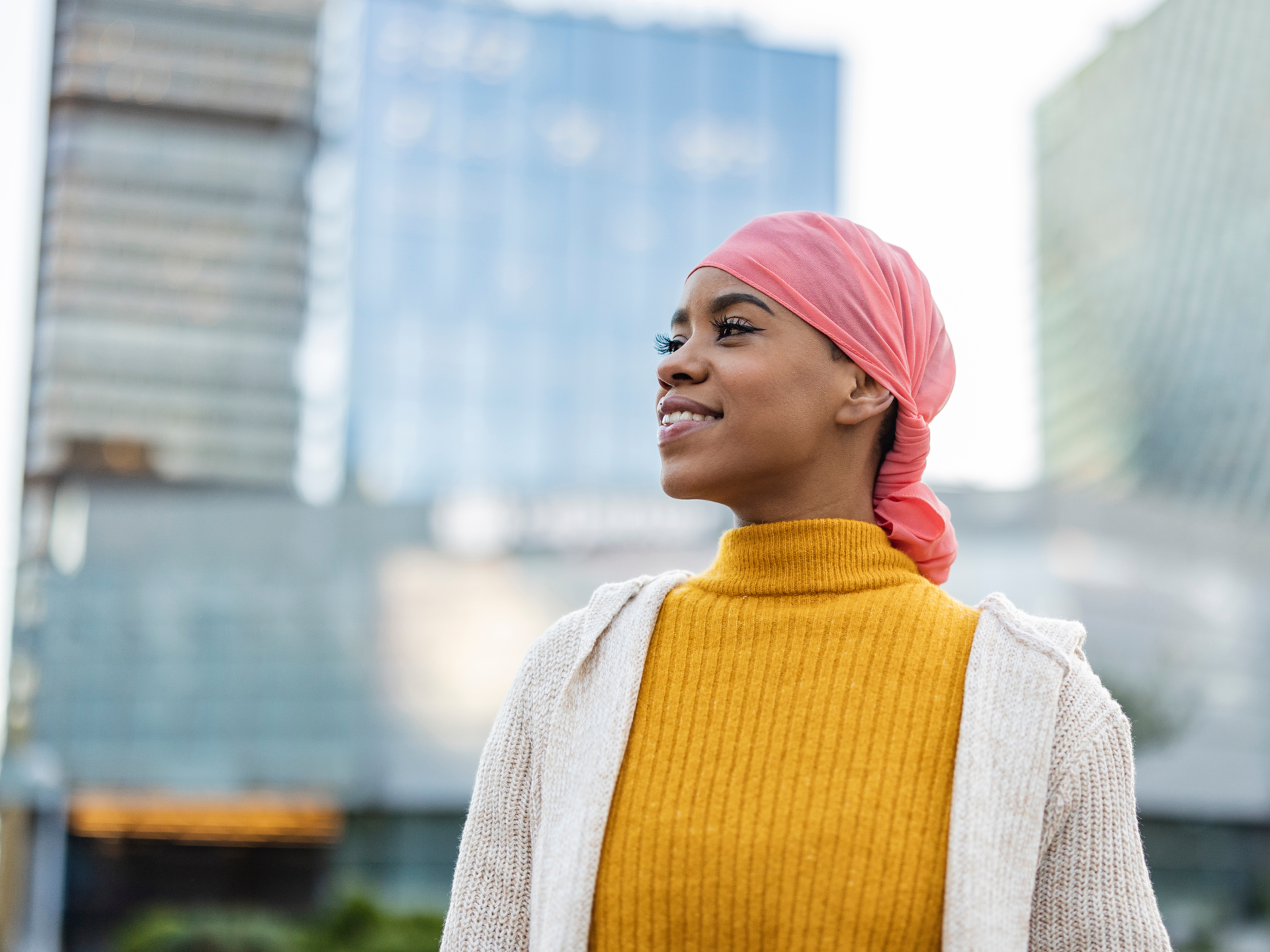 A person wearing a pink headscarf and a yellow turtleneck sweater smiles while standing outdoors in an urban area with tall glass buildings in the background.