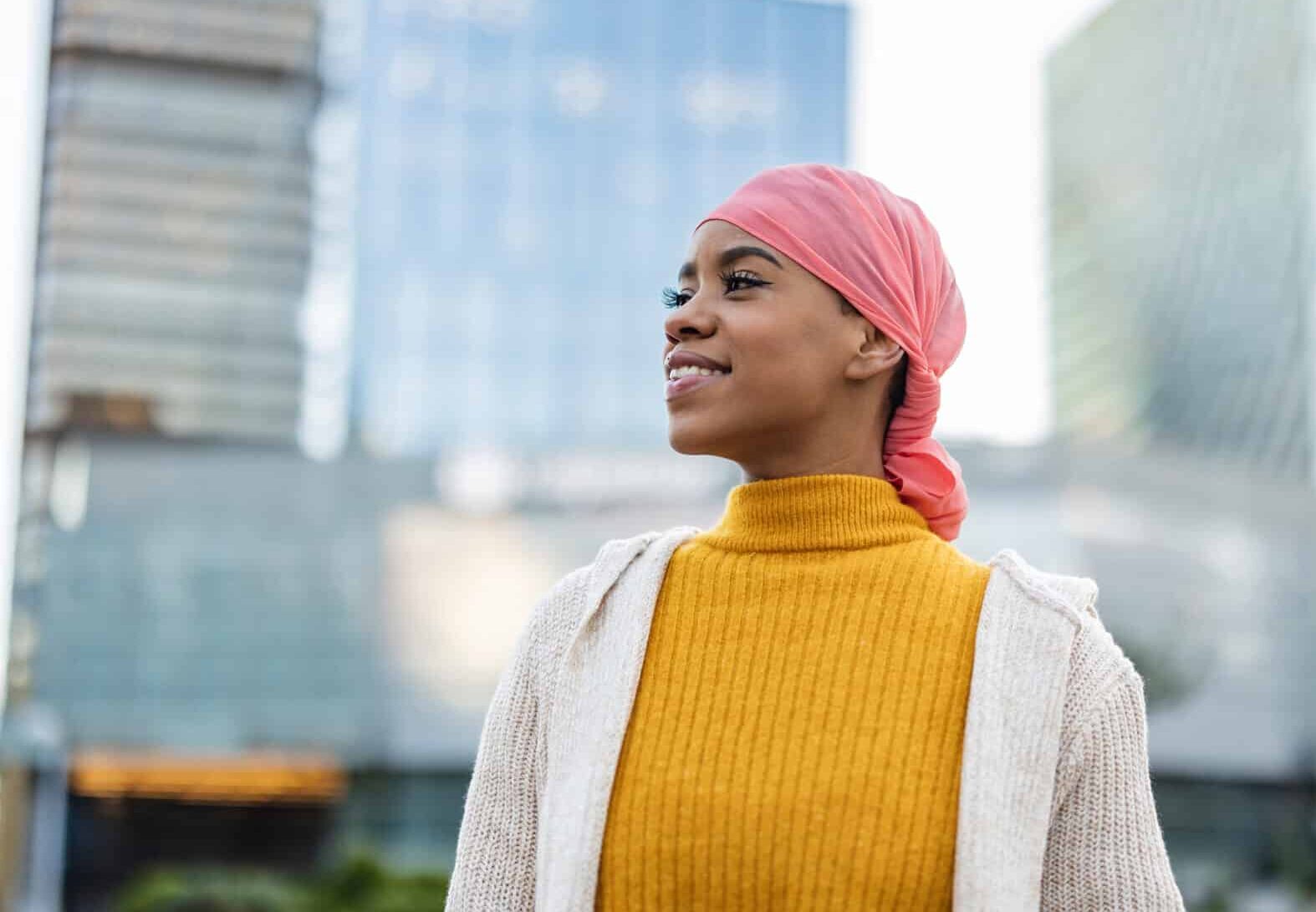 A person wearing a pink headscarf and a yellow turtleneck smiles while standing outdoors in a cityscape with tall buildings in the background.