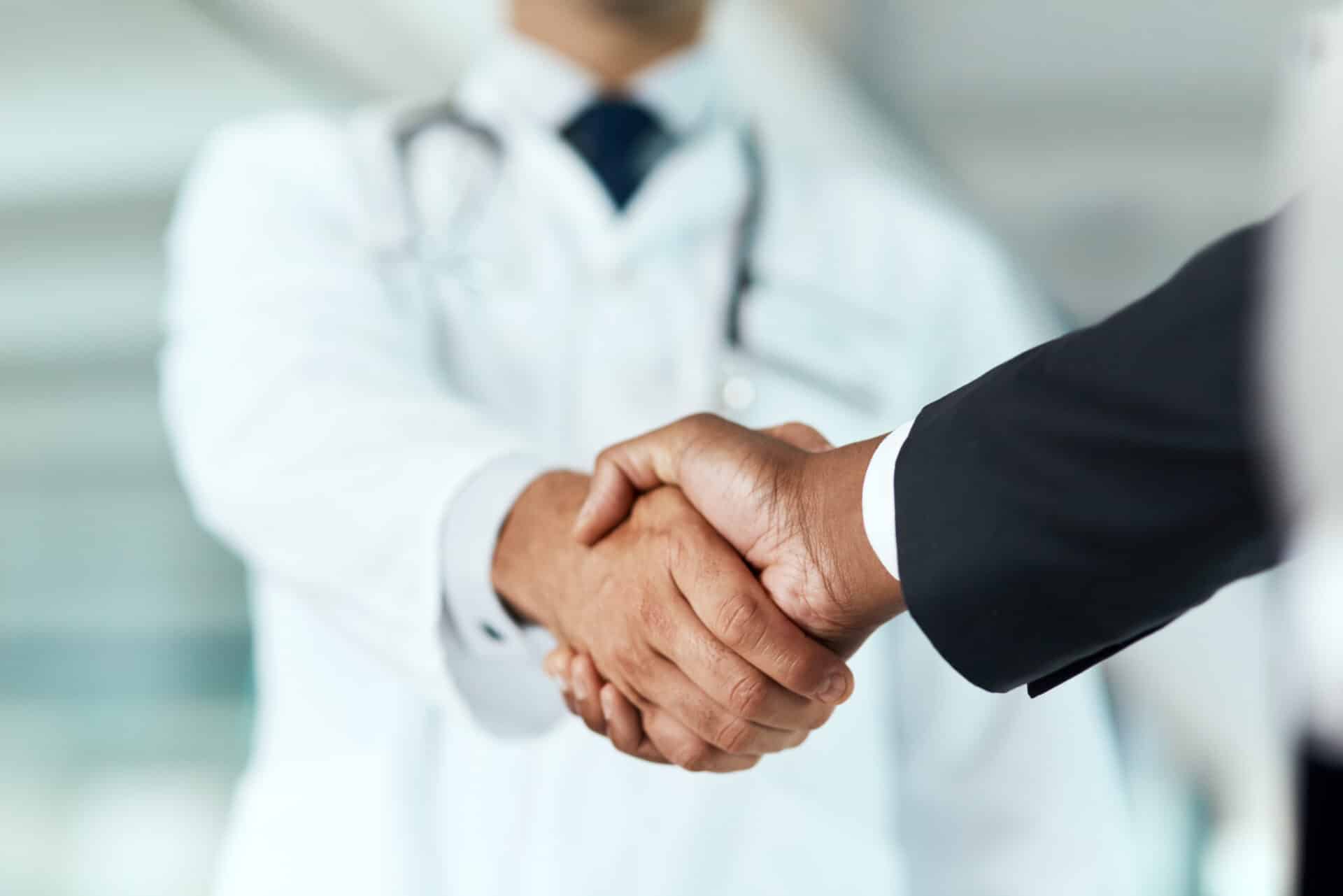 A doctor in a white coat shakes hands with a person in a business suit, symbolizing a partnership or agreement. The background is blurred, highlighting the handshake as the focus of the image.