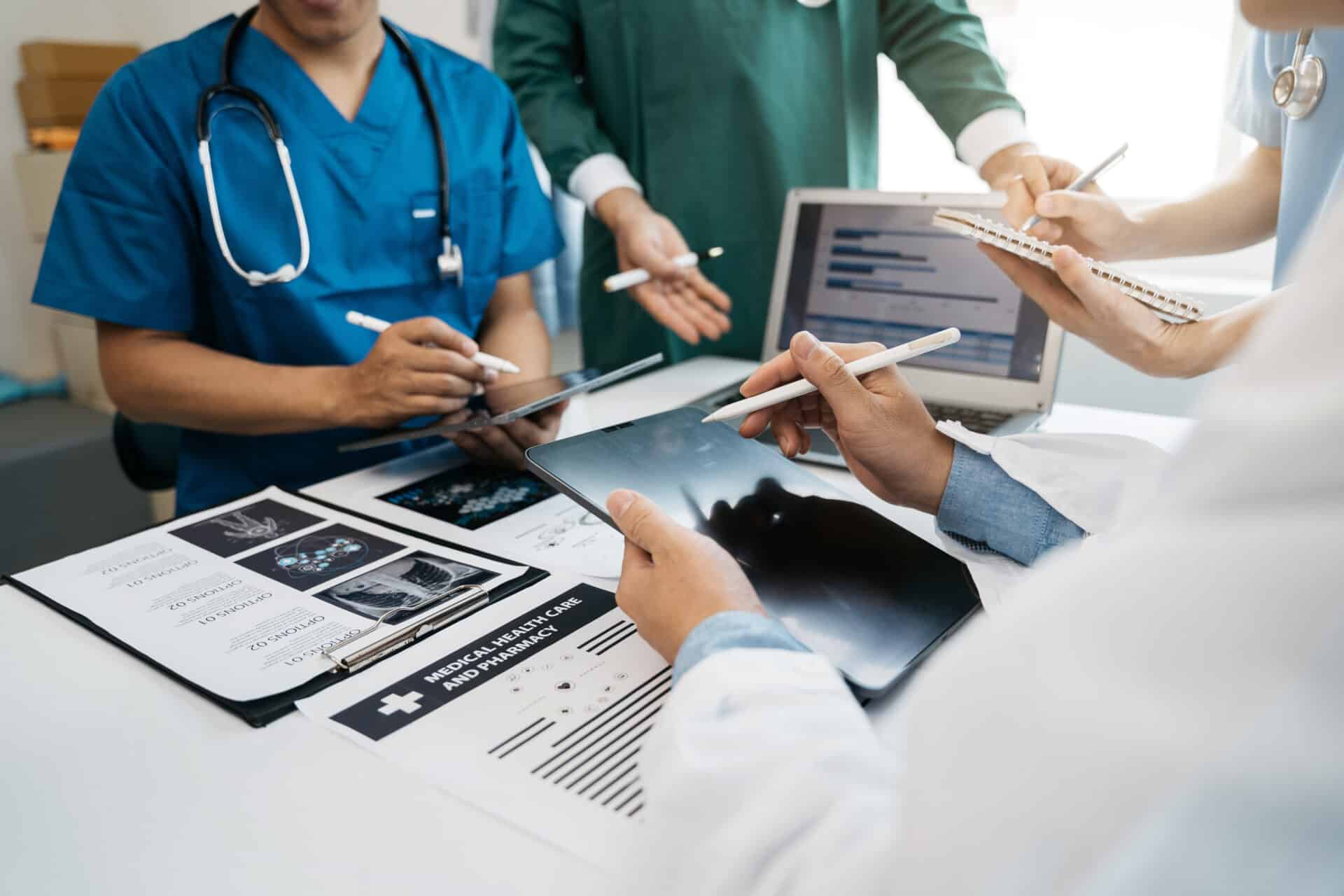 A group of healthcare professionals discusses medical images displayed on a tablet. Laptops and documents with charts are on the table, including X-ray images and medical documents. They are wearing medical scrubs and lab coats.