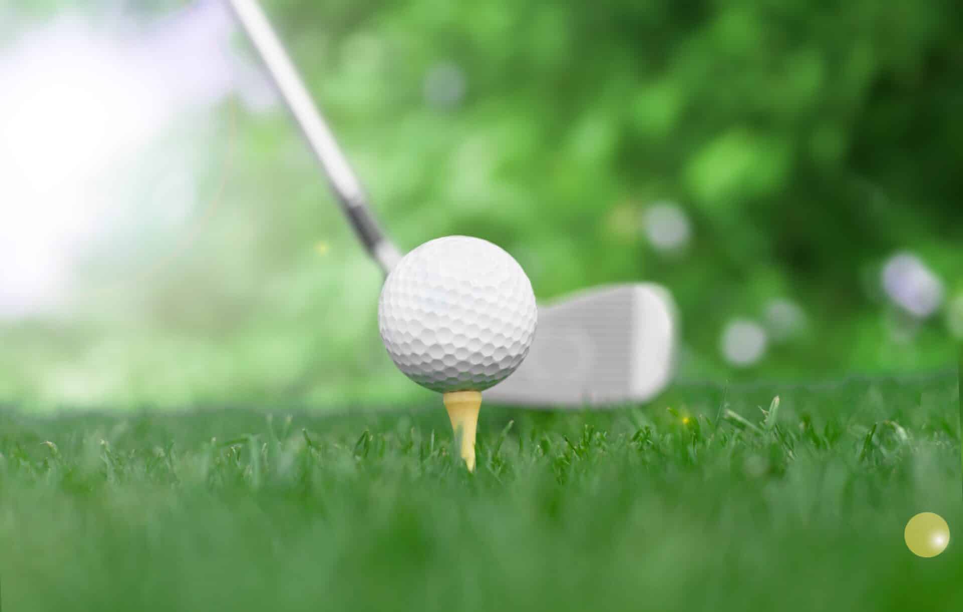 A close-up of a golf ball on a tee with a golf club poised to hit it. The background is a blurred green field, suggesting a golf course setting. Sunlight softly illuminates the scene.