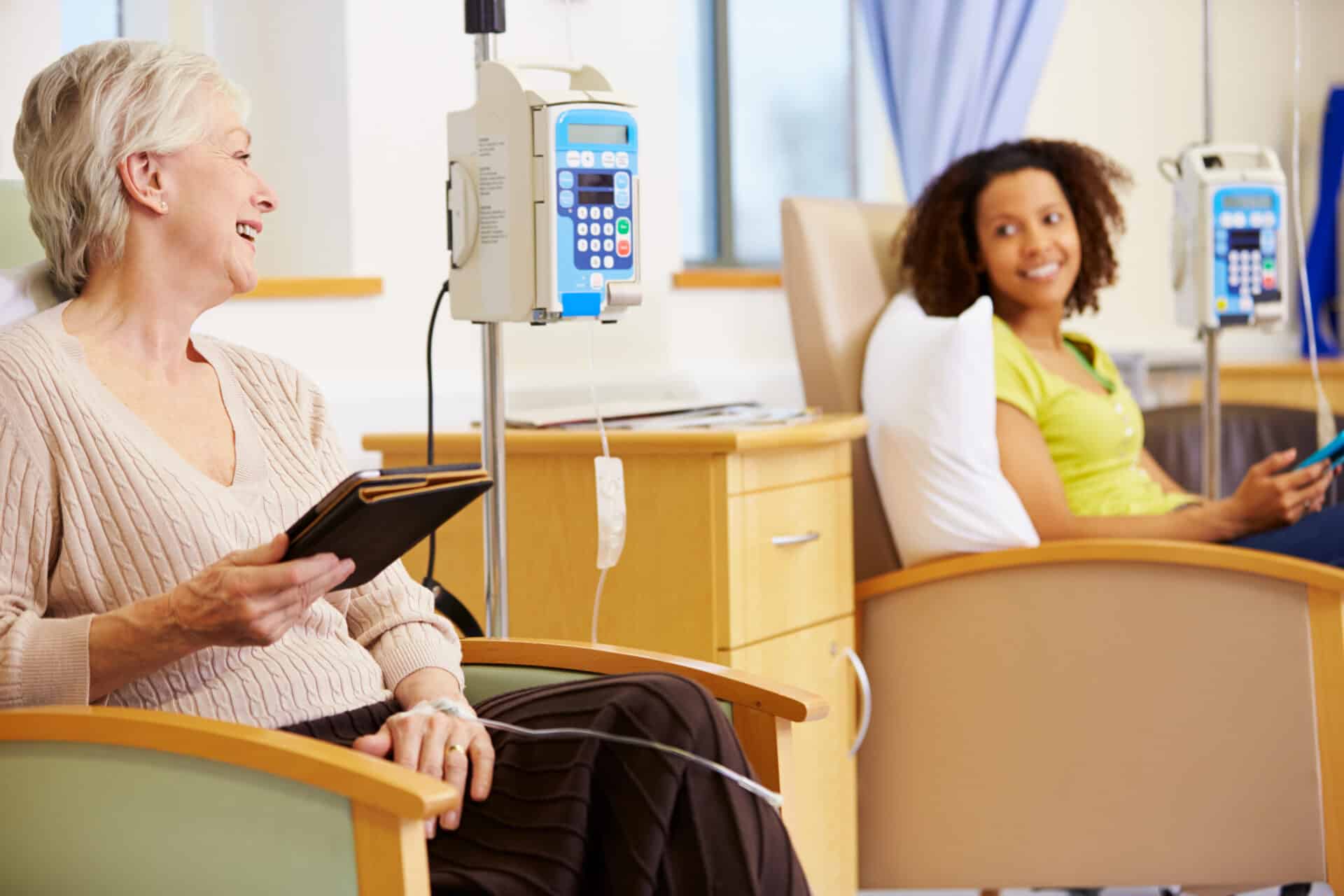 Two women receiving IV therapy sit in medical chairs in a hospital room. The older woman in the foreground holds a tablet and smiles, while the younger woman in the background looks over, smiling with a phone in her hand.