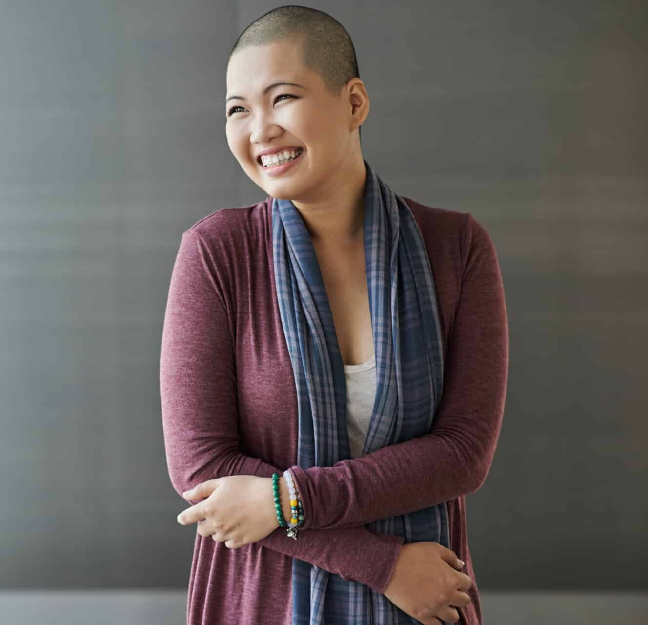 A person with a shaved head smiles, wearing a long-sleeved maroon shirt, white top, and a blue plaid scarf. They have their arms crossed and are wearing bracelets. The background is a muted gray.