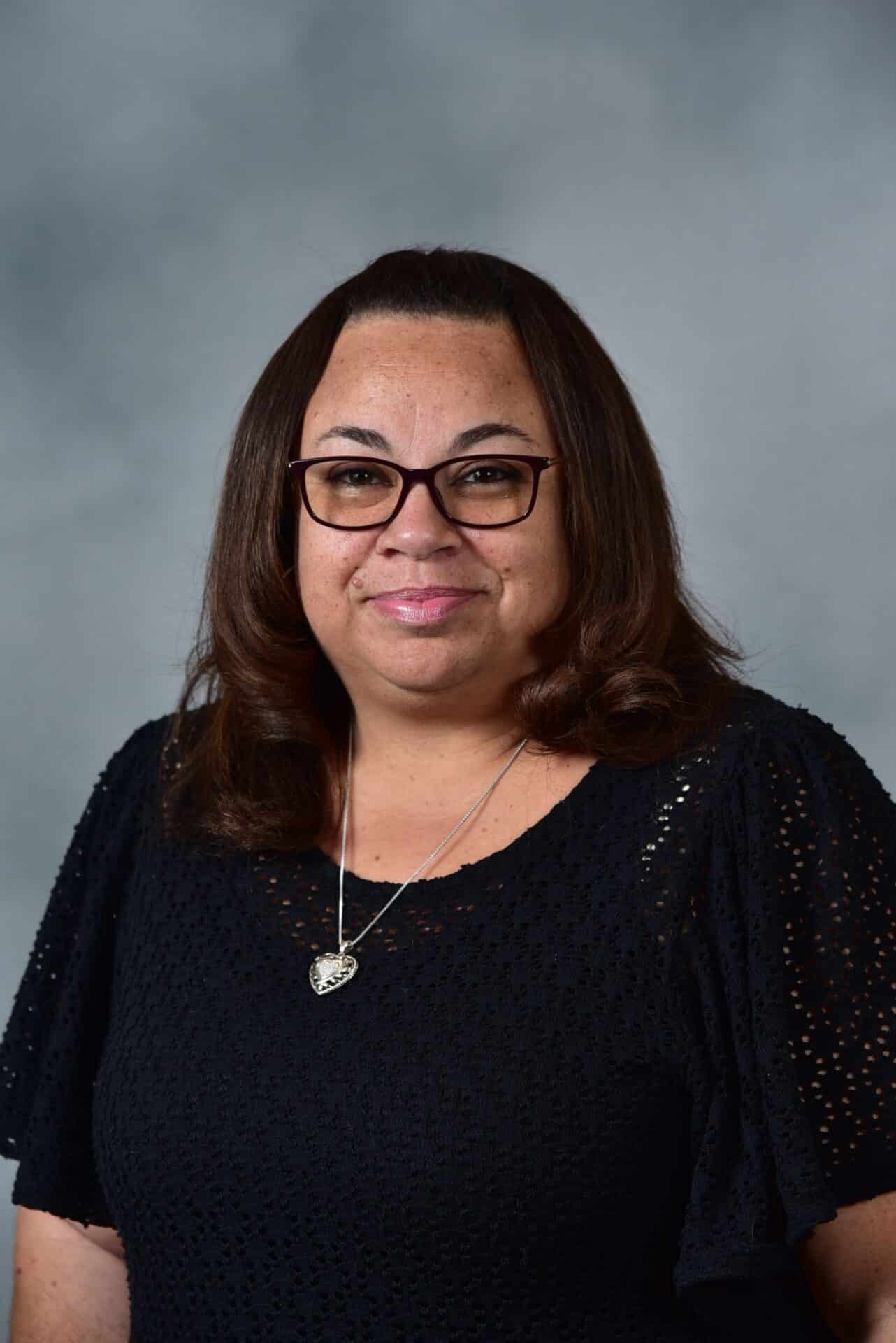 A woman with shoulder-length brown hair and glasses, wearing a black knitted top and a heart-shaped necklace, smiles in front of a gray background.