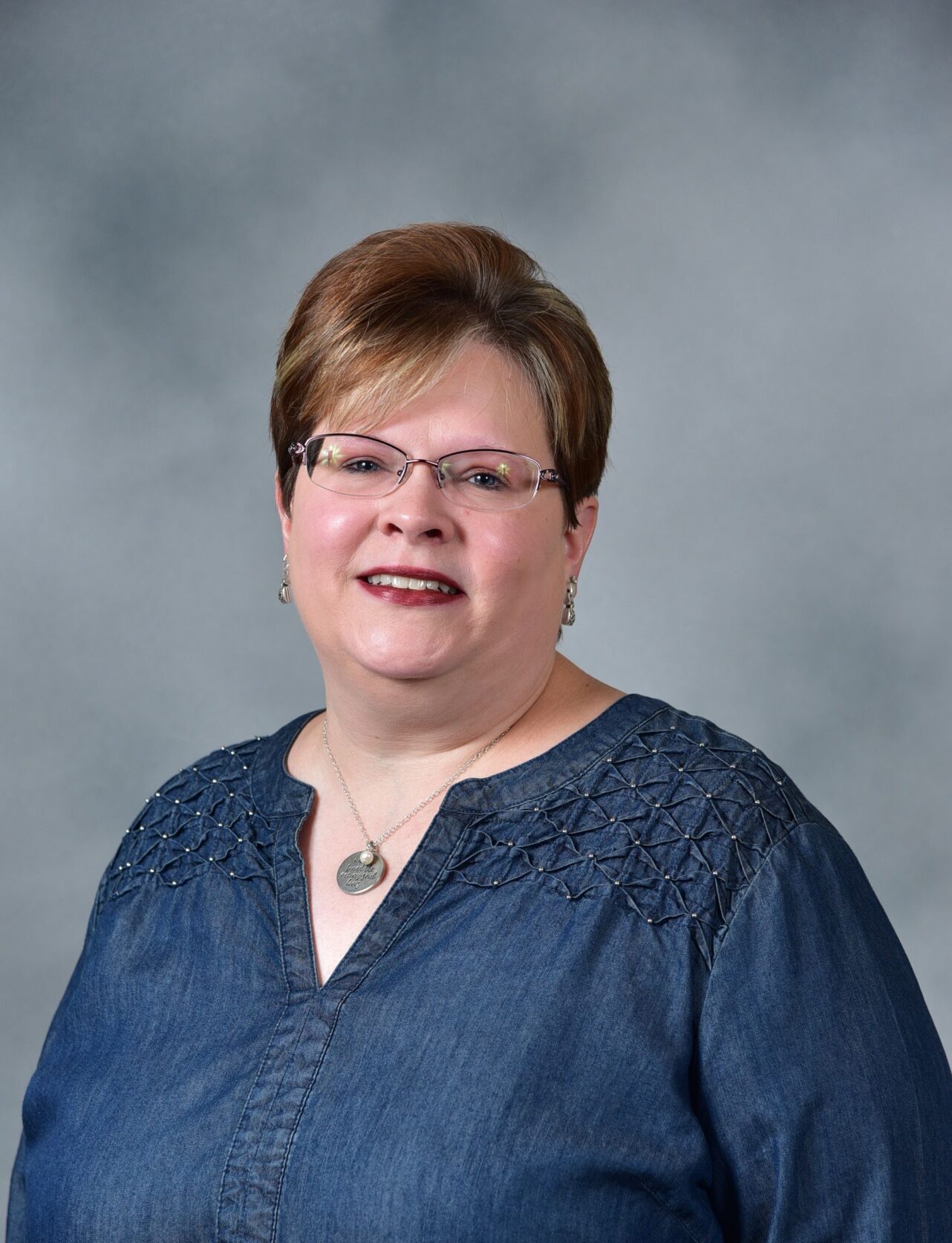 A person wearing glasses and a blue blouse with intricate patterns on the shoulders. They have short hair and are smiling at the camera, standing against a plain gray background.