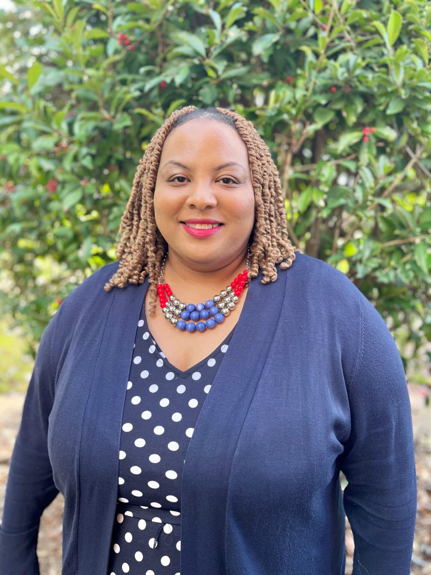 A person with curly hair stands outdoors in front of a leafy green background. They are wearing a navy blue cardigan, a polka dot dress, and a colorful beaded necklace, smiling at the camera.