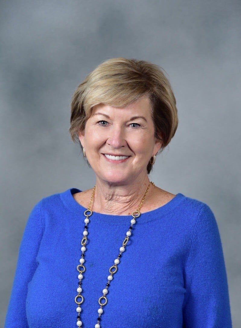 A woman with short blond hair smiles at the camera. She is wearing a blue sweater and a beaded necklace. The background is plain gray.