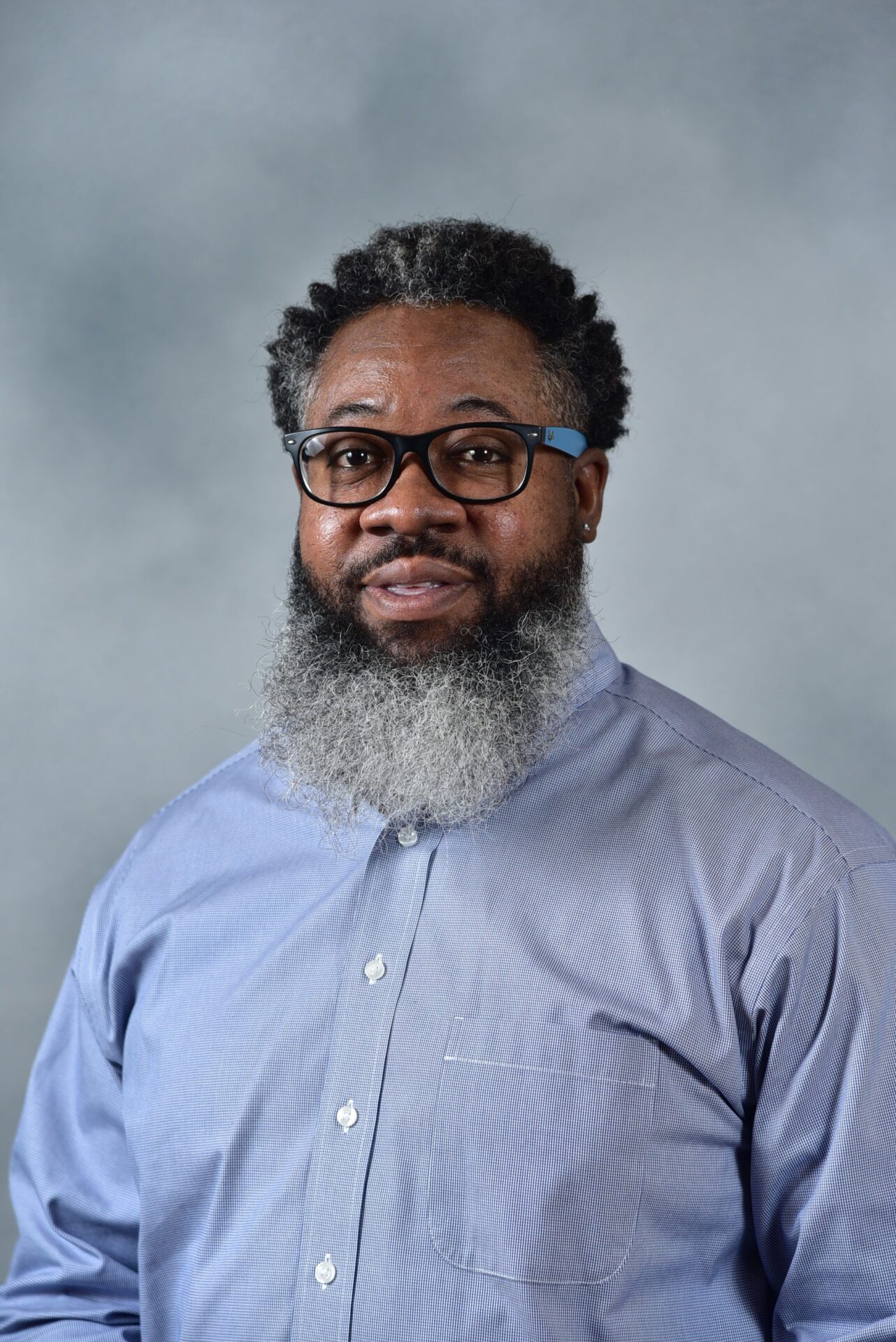 A man with glasses, a long gray beard, and short curly hair is wearing a light blue button-up shirt. He is standing against a neutral gray background, looking at the camera with a slight smile.