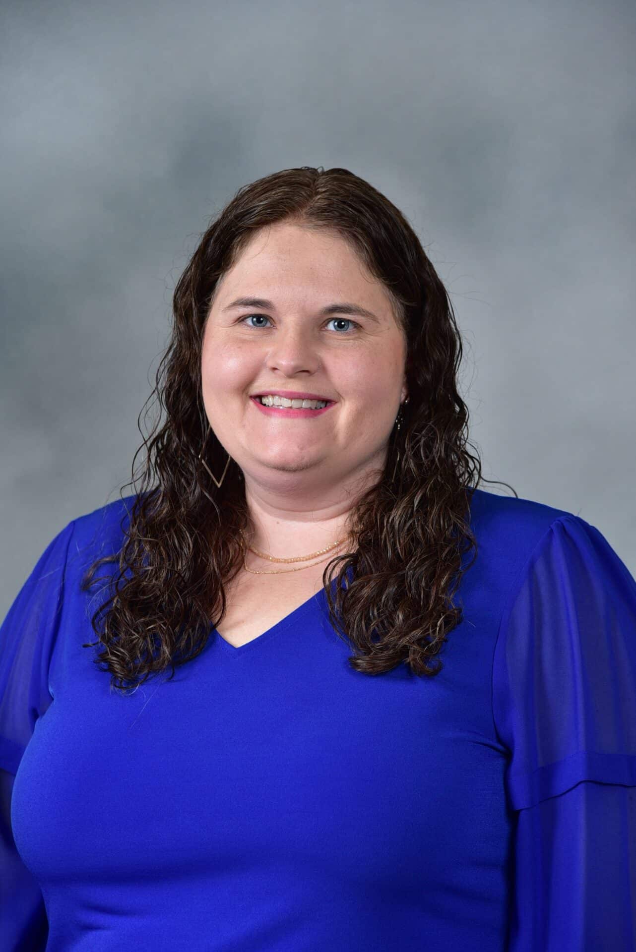 A woman with long brown curly hair wearing a royal blue top smiles at the camera against a gray background.