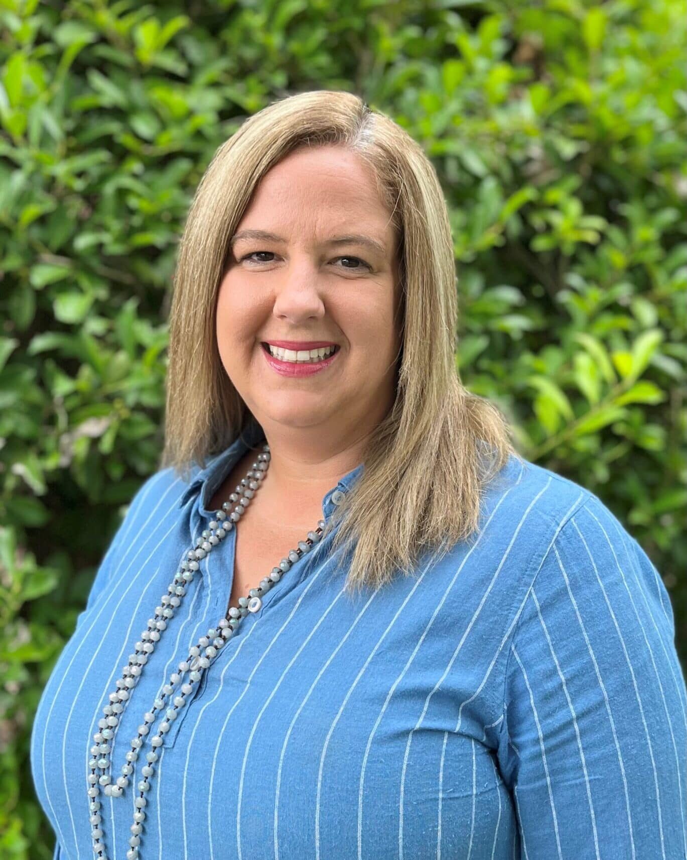 A smiling woman with shoulder-length blonde hair is wearing a blue pinstripe blouse and a pearl necklace. She is standing in front of a lush green leafy background.