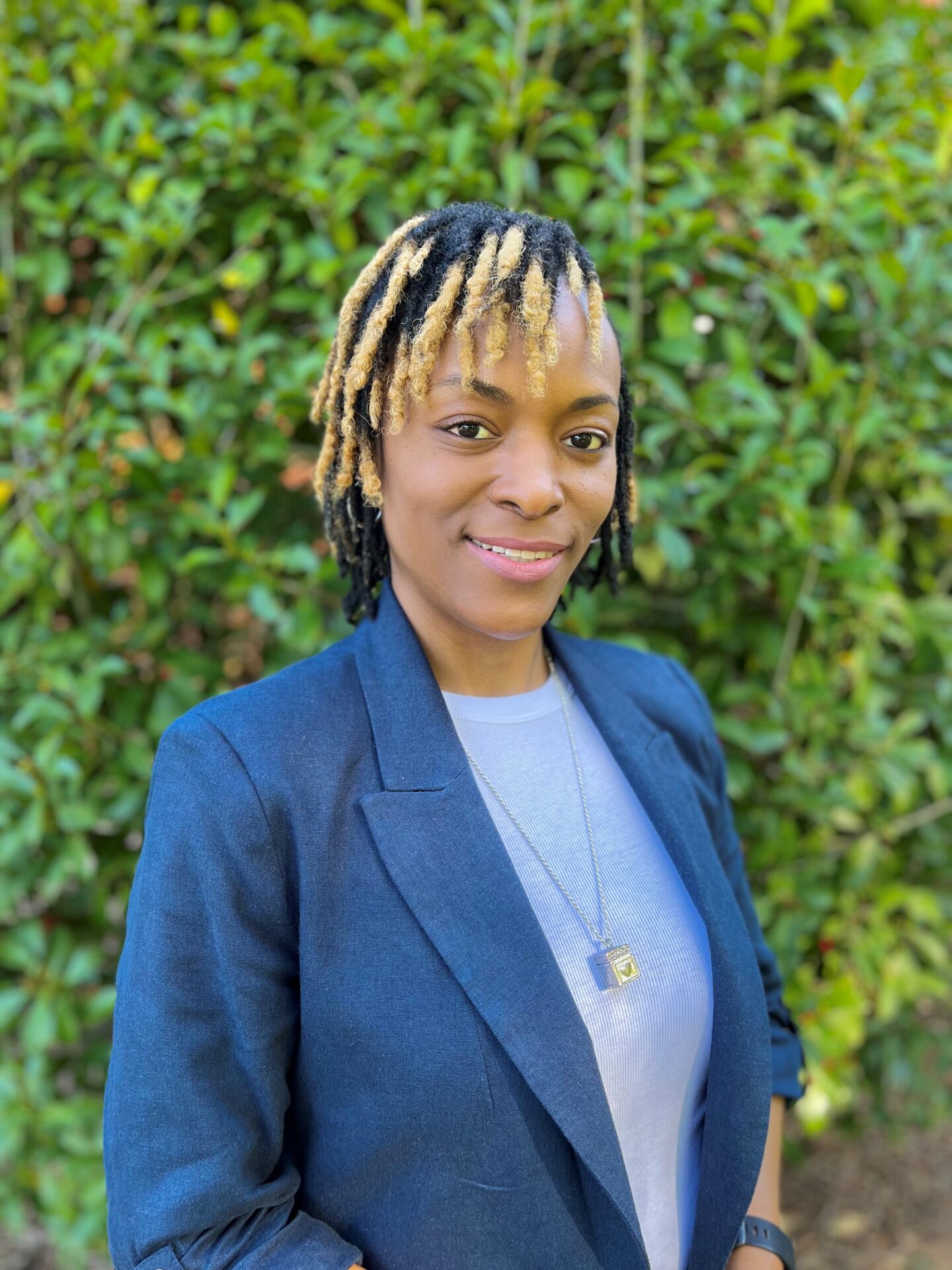 A person wearing a blue blazer and a light-colored shirt with a pendant necklace is smiling while standing in front of a lush green leafy background.