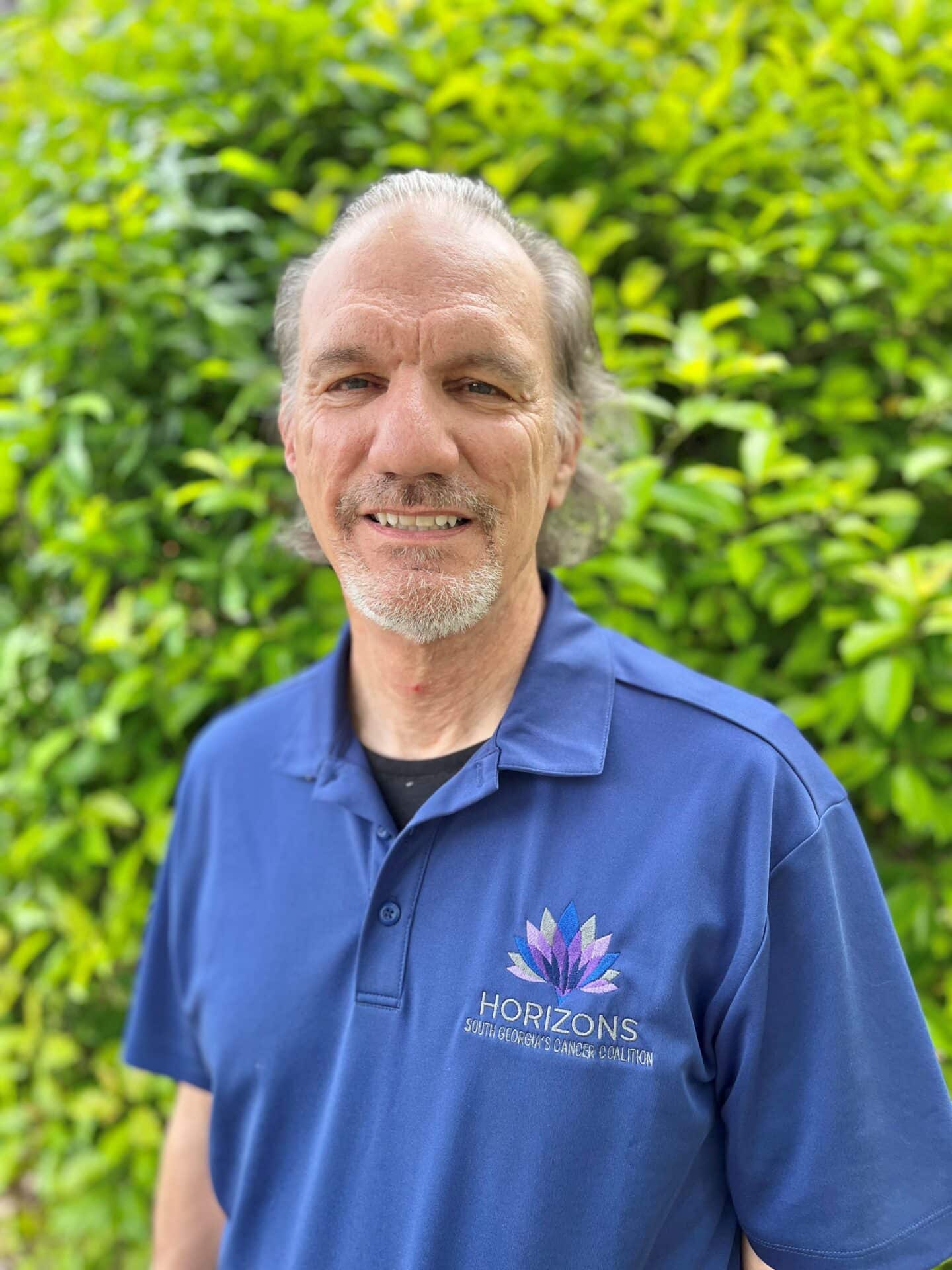 A man with a receding hairline smiles while standing in front of lush green foliage. He is wearing a blue polo shirt with Horizons and South Georgia's Cancer Coalition embroidered on it.