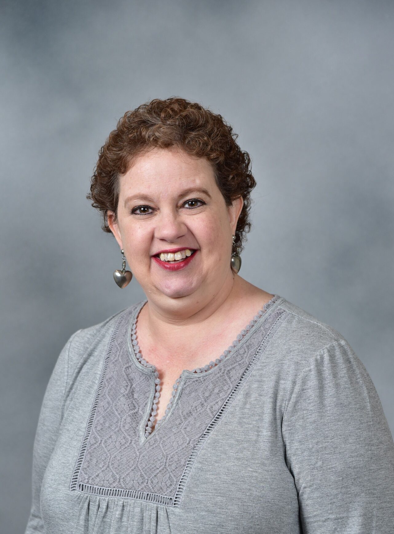 A person with curly brown hair, wearing a gray top with lace detailing and heart-shaped earrings, smiles against a gray background.