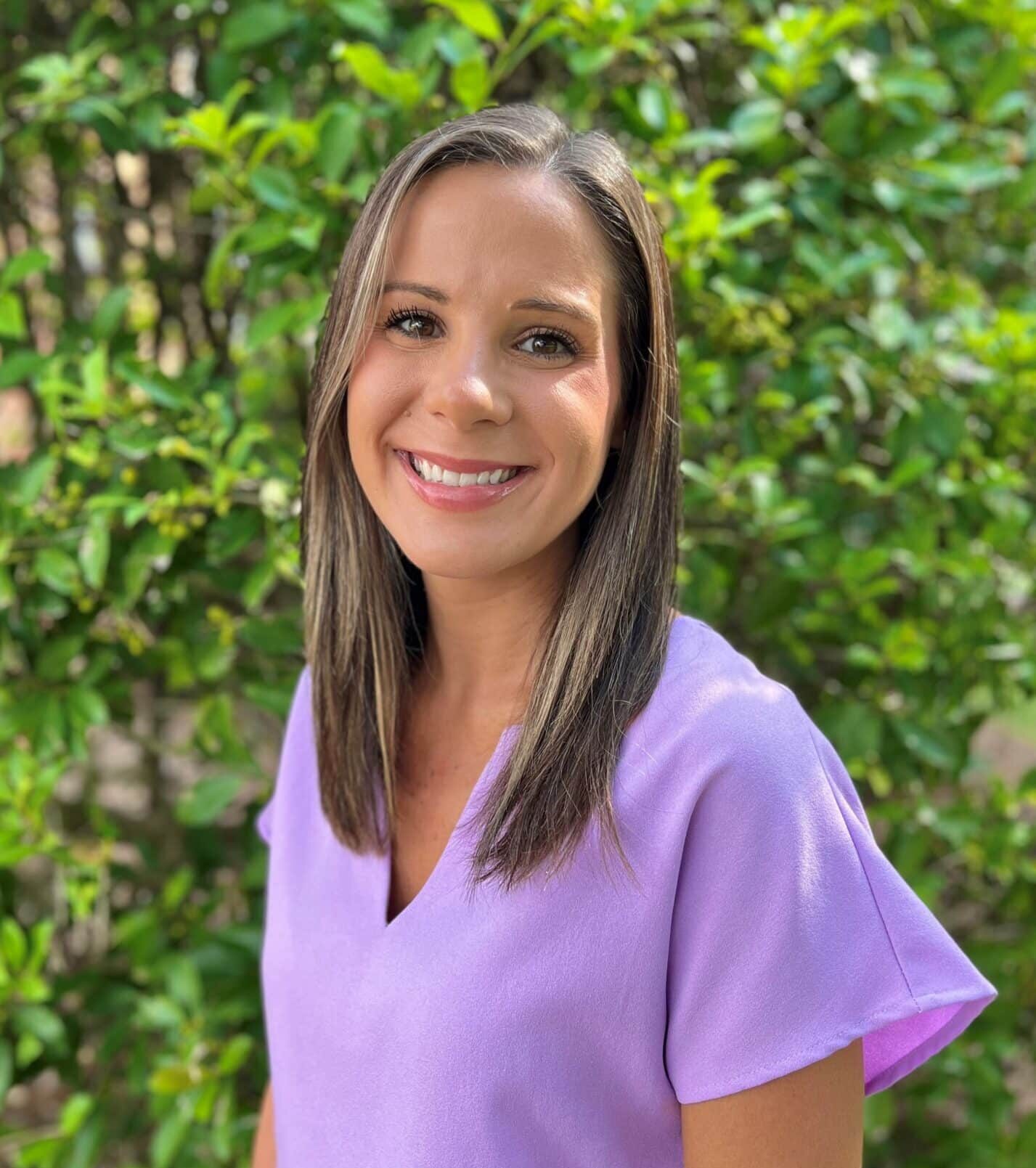 A woman with straight hair wearing a lavender blouse smiles while standing in front of green leafy bushes.