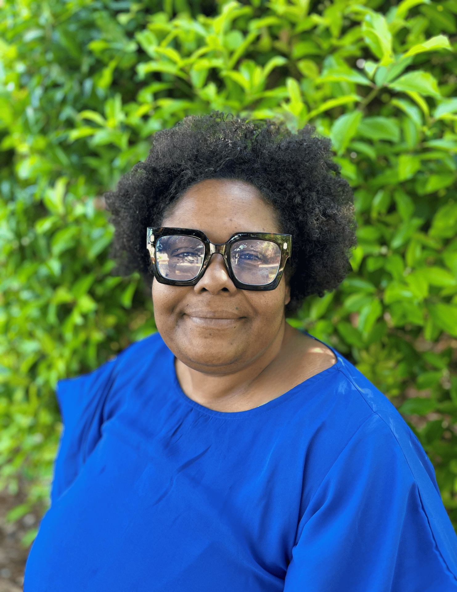 A person wearing large glasses and a bright blue shirt smiles while standing in front of a lush green bush.