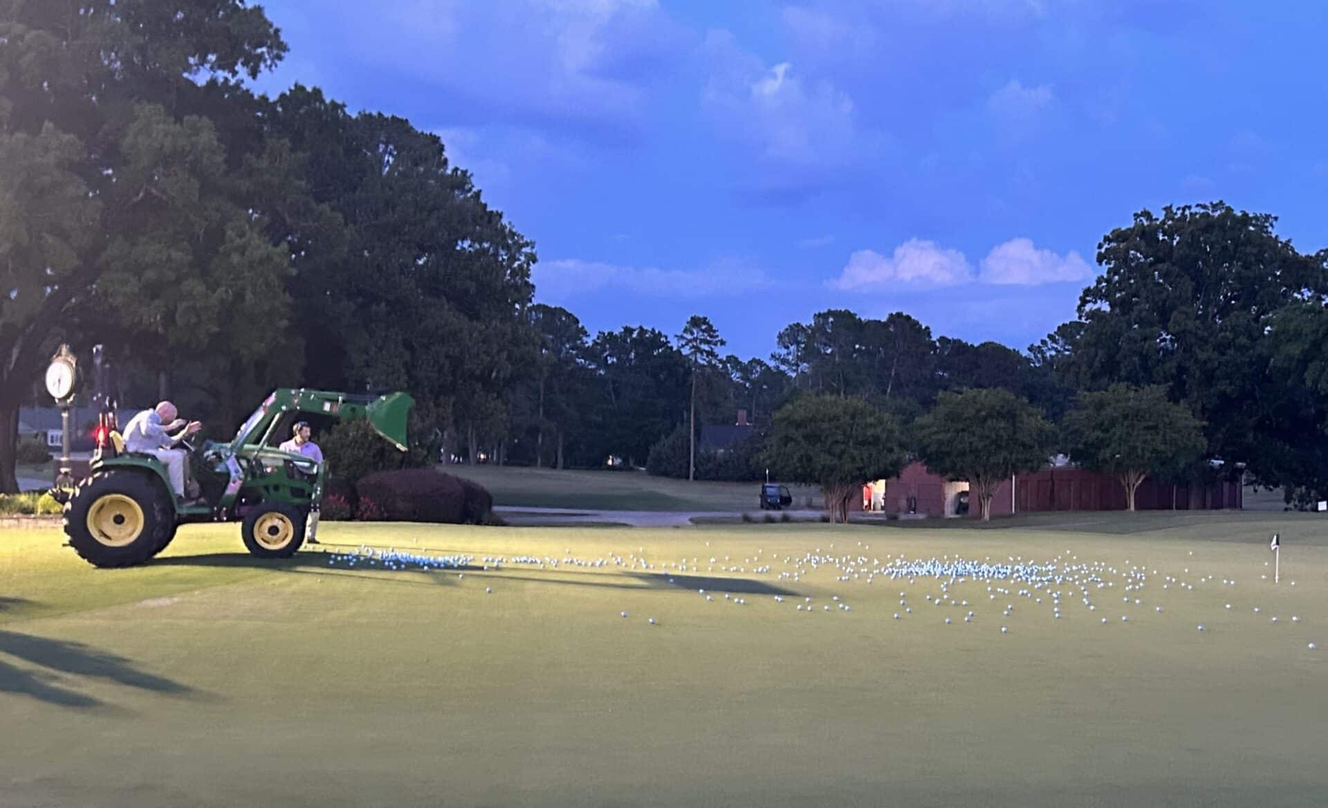 A person operates a small green tractor on a golf course in the evening. The tractor's bucket is raised, spilling numerous white golf balls onto the green. Trees and a small building are visible in the background under a blue sky.