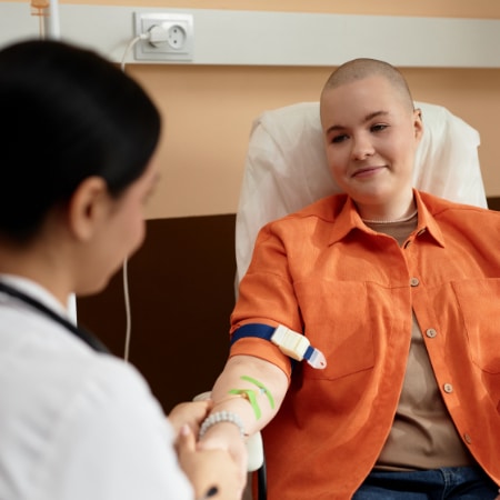 A smiling person with a shaved head sits in a clinic chair, wearing an orange shirt, as a healthcare worker prepares their arm.